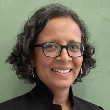 Head shot of Sanchita Balachandran wearing glasses and a dark jacket with pale green background