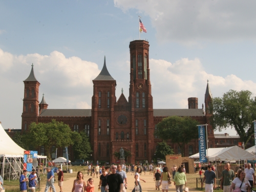 Red brick castle with sandy walkways in front, people crossing the paths, and white tents in foreground.