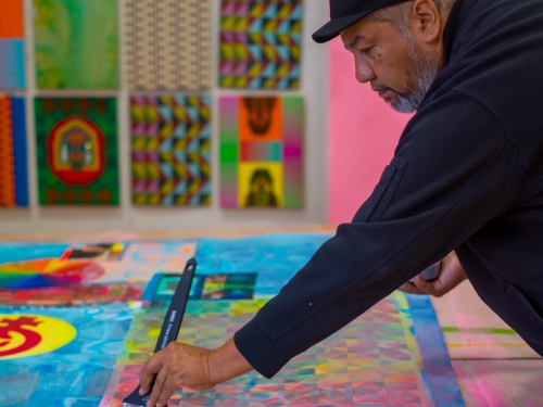 Man in black long sleeves and black cap leans over colorful tapesty, one arm out holding a paintbrush.